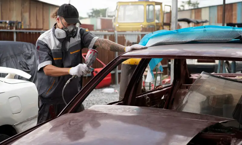 A mechanic in protective gear uses a spray gun to paint the roof of a car in a busy auto repair shop with various vehicles around A mechanic in protective gear uses a spray gun to paint the roof of a car in a busy auto repair shop with various vehicles around